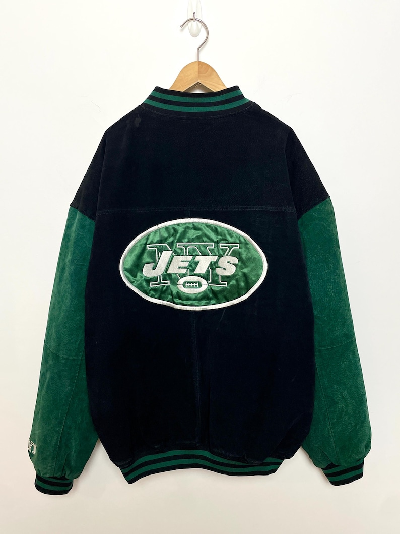 Pode incluir: Casaco universit&aacute;rio vintage dos Jets, preto e verde. O casaco tem mangas verdes e um log&oacute;tipo dos Jets verde e branco nas costas. O casaco est&aacute; pendurado num cabide de madeira.