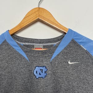 Puede incluir: Una camiseta deportiva gris con detalles en azul claro y un logotipo azul claro. La camiseta presenta un logotipo de Nike y un logotipo en el pecho. La camiseta est&aacute; colgada en una percha de madera.