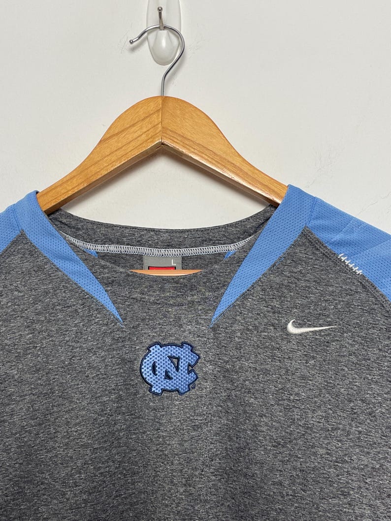 Puede incluir: Una camiseta deportiva gris con detalles en azul claro y un logotipo azul claro. La camiseta presenta un logotipo de Nike y un logotipo en el pecho. La camiseta est&aacute; colgada en una percha de madera.