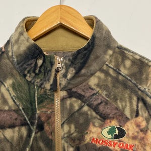 May include: A camouflage fleece jacket with a full zipper and a stand-up collar. The jacket features the "Mossy Oak" logo embroidered in red and green. The jacket is hanging on a wooden hanger.