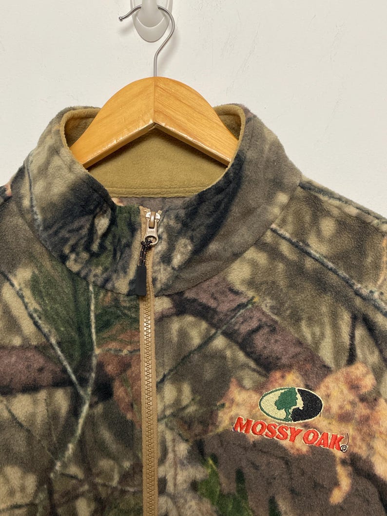 May include: A camouflage fleece jacket with a full zipper and a stand-up collar. The jacket features the "Mossy Oak" logo embroidered in red and green. The jacket is hanging on a wooden hanger.