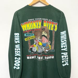 May include: Green long-sleeve t-shirt with a cartoon illustration of a man riding a motorcycle. The shirt features the text "Whiskey Pete's Tavern & Eatery" and "Bike Week 2002".