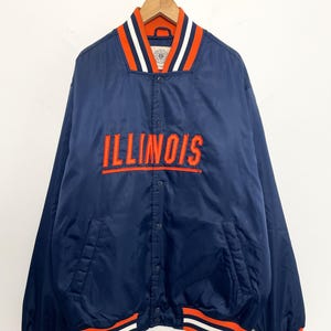Puede incluir: Chaqueta de satén azul marino con la palabra "ILLINOIS" en letras naranjas en el pecho. La chaqueta tiene ribetes a rayas naranjas y blancas en el cuello, los puños y el dobladillo. Cuelga de una percha de madera.