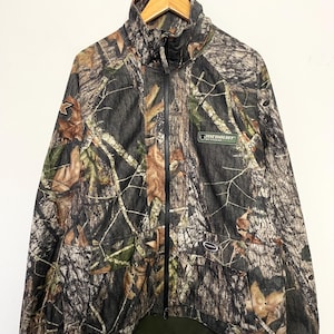 Pode incluir: Casaco camuflado com z&iacute;per completo, gola alta e esquema de cores verde e preto. O casaco apresenta um padr&atilde;o de floresta com detalhes de folhas marrons e verdes. Um pequeno patch verde com texto branco &eacute; vis&iacute;vel no peito.