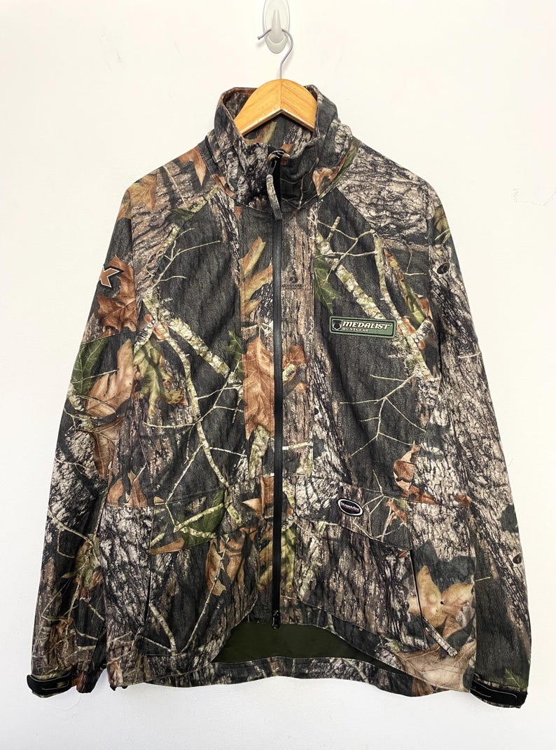 Pode incluir: Casaco camuflado com z&iacute;per completo, gola alta e esquema de cores verde e preto. O casaco apresenta um padr&atilde;o de floresta com detalhes de folhas marrons e verdes. Um pequeno patch verde com texto branco &eacute; vis&iacute;vel no peito.