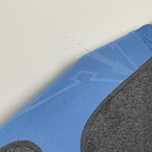 Puede incluir: Primer plano de una camiseta deportiva azul con un patr&oacute;n de rayo y un panel gris. La camiseta tiene una textura perforada y una costura curva. La tela parece ser un material ligero y transpirable.
