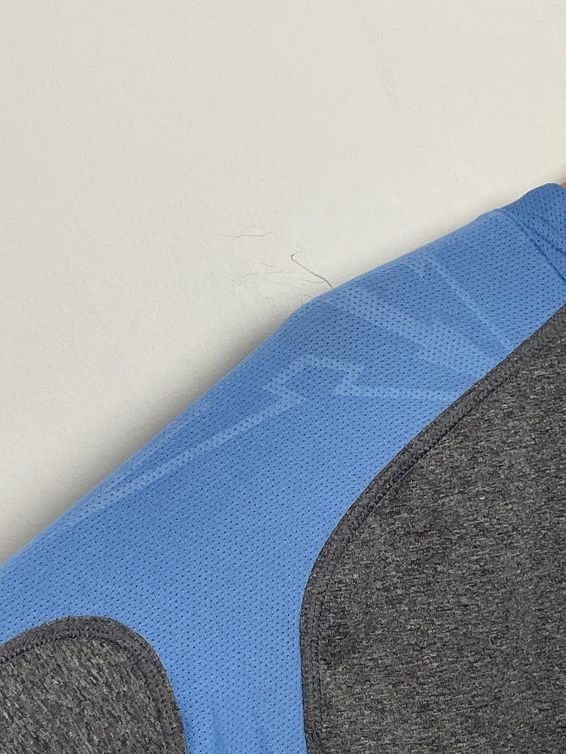 Puede incluir: Primer plano de una camiseta deportiva azul con un patr&oacute;n de rayo y un panel gris. La camiseta tiene una textura perforada y una costura curva. La tela parece ser un material ligero y transpirable.