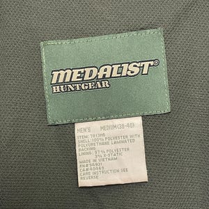 Pode incluir: Close-up de um colete de ca&ccedil;a verde com uma etiqueta. A etiqueta diz "MEDALIST HUNTGEAR" em letras brancas. Abaixo, uma etiqueta com o tamanho "MEDIUM (38-40)" e a composi&ccedil;&atilde;o do material.