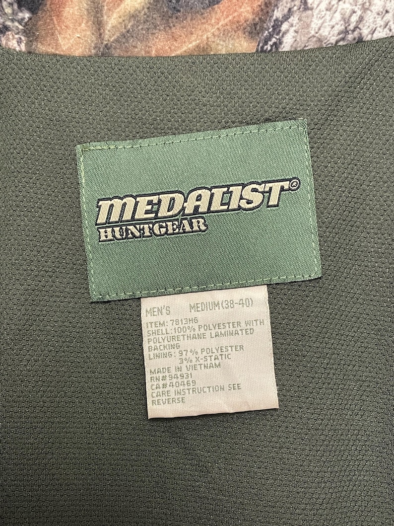 Pode incluir: Close-up de um colete de ca&ccedil;a verde com uma etiqueta. A etiqueta diz "MEDALIST HUNTGEAR" em letras brancas. Abaixo, uma etiqueta com o tamanho "MEDIUM (38-40)" e a composi&ccedil;&atilde;o do material.