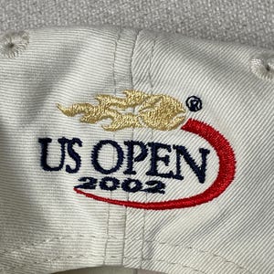 May include: A cream-colored baseball cap featuring an embroidered logo with the text US OPEN 2002 in dark blue, a gold flame-like graphic above the text, and a red curved swoosh accenting the bottom of the lettering on the front panel.