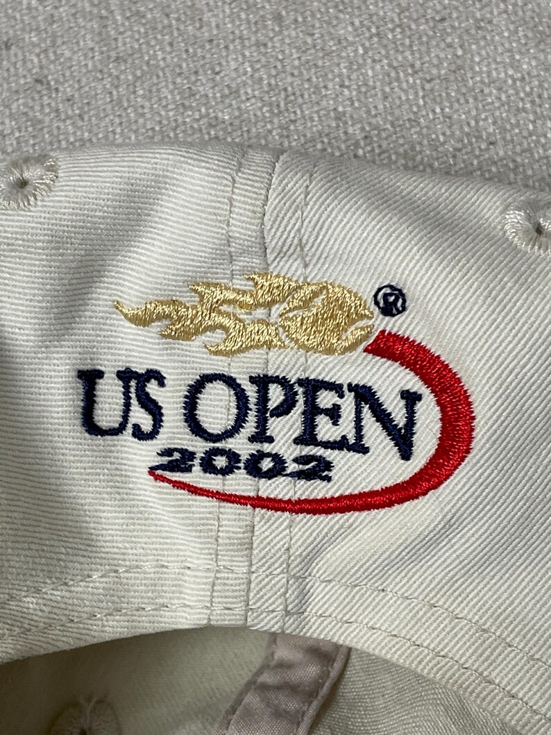May include: A cream-colored baseball cap featuring an embroidered logo with the text US OPEN 2002 in dark blue, a gold flame-like graphic above the text, and a red curved swoosh accenting the bottom of the lettering on the front panel.