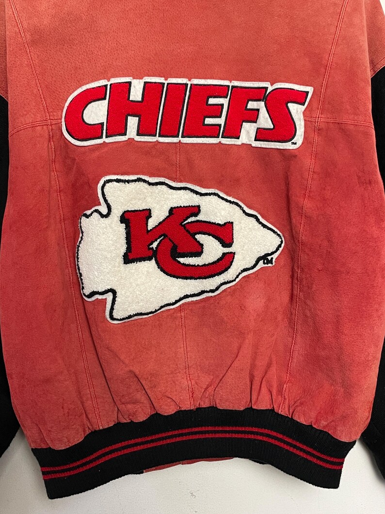 Puede incluir: Chaqueta de cuero roja con cuello y pu&ntilde;os a rayas negras y rojas. La parte posterior de la chaqueta presenta un gran logotipo "CHIEFS" blanco y rojo con un logotipo "KC" blanco y rojo m&aacute;s peque&ntilde;o debajo.