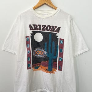 May include: White t-shirt with a graphic print of a cactus and a moon. The text "Arizona Super Bowl" is printed vertically on the sides of the graphic. The graphic also includes the text "Super XXX Bowl".