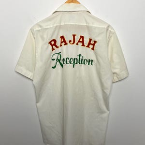 May include: Cream-colored short-sleeve shirt with the words "RAJAH Reception" embroidered on the back in red and green. The shirt is hanging on a white wall.