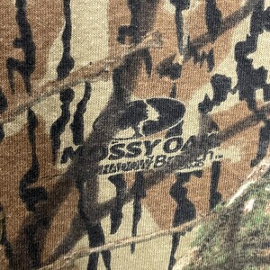 May include: Close-up of a camouflage pattern in shades of brown, tan, and black. The fabric has a textured appearance. The Mossy Oak logo is visible in the centre of the image.