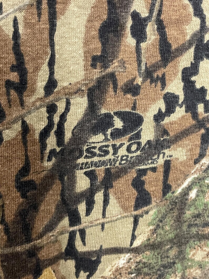 May include: Close-up of a camouflage pattern in shades of brown, tan, and black. The fabric has a textured appearance. The Mossy Oak logo is visible in the centre of the image.