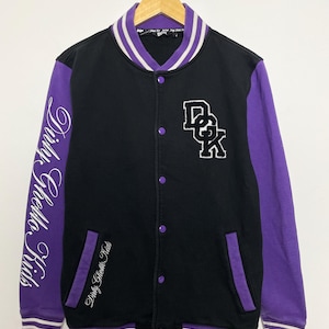 以下が含まれることがあります： 胸に「DGK」の文字が入った黒と紫のスタジアムジャンパー。袖は紫で、白の筆記体で「Dirty Ghetto Kids」と書かれています。ジャケットにはストライプの襟と袖口が付いています。