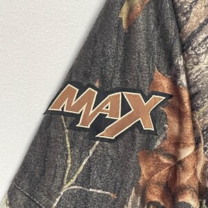 Pode incluir: Tecido camuflado com a palavra "MAX" em letras marrons e brancas. O tecido tem uma mistura de tons marrons, verdes e cinzas, que se assemelham a um ambiente florestal. O patch "MAX" &eacute; delineado em branco e sobre um fundo preto.