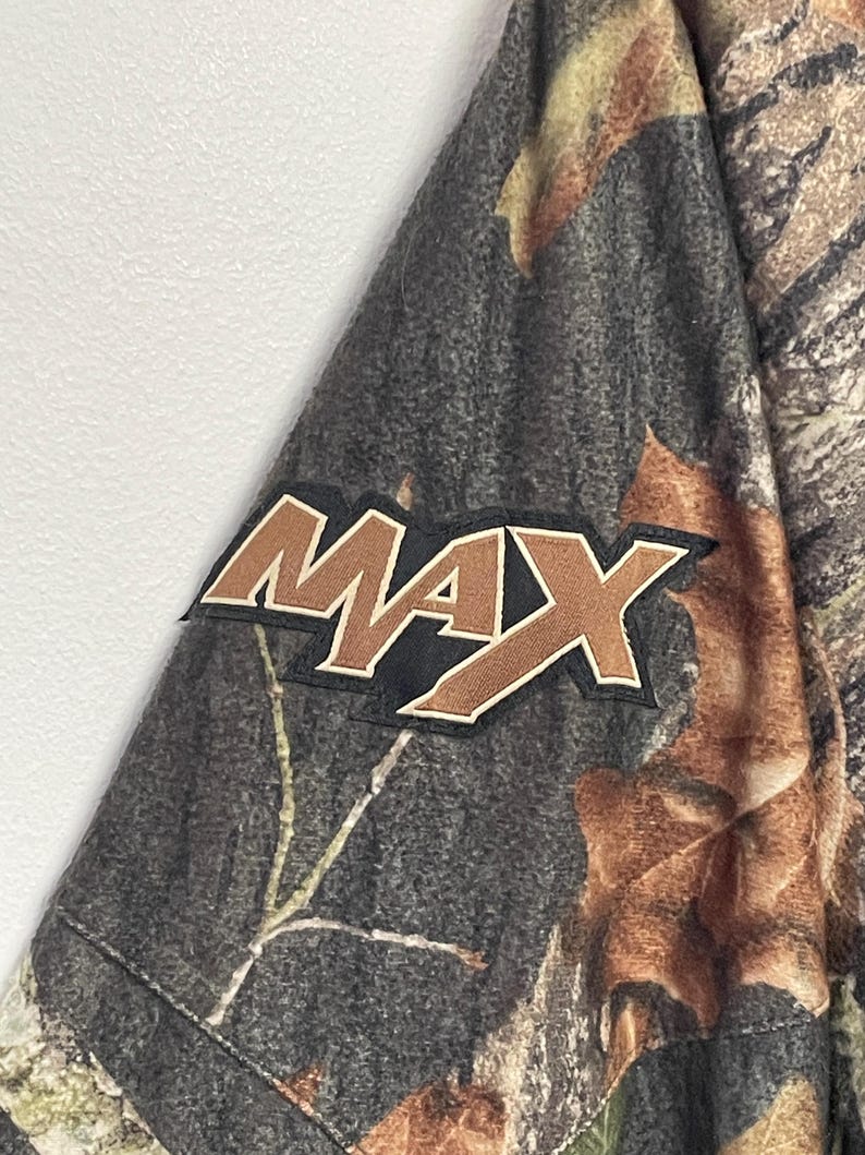 Pode incluir: Tecido camuflado com a palavra "MAX" em letras marrons e brancas. O tecido tem uma mistura de tons marrons, verdes e cinzas, que se assemelham a um ambiente florestal. O patch "MAX" &eacute; delineado em branco e sobre um fundo preto.