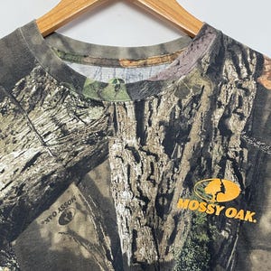 May include: A camouflage-patterned t-shirt with a crew neck, hanging on a wooden hanger. The shirt features a brown, green, and tan tree bark design. The "Mossy Oak" logo is printed in orange on the right side of the shirt.