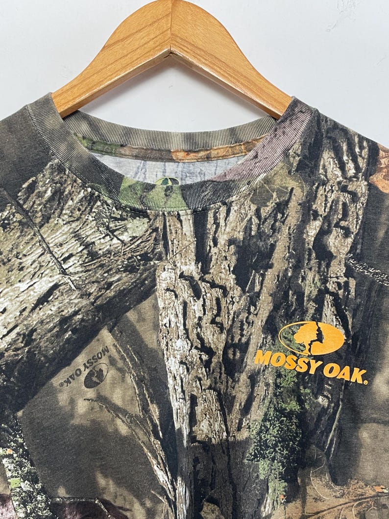 May include: A camouflage-patterned t-shirt with a crew neck, hanging on a wooden hanger. The shirt features a brown, green, and tan tree bark design. The "Mossy Oak" logo is printed in orange on the right side of the shirt.