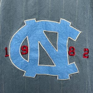 May include: A dark blue pinstriped garment featuring a large, light blue embroidered NC logo in the center. The numbers 19 and 82 are stitched in red on either side of the logo, with 19 on the left and 82 on the right, all on a textured fabric.