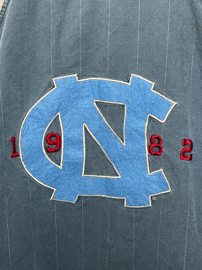 May include: A dark blue pinstriped garment featuring a large, light blue embroidered NC logo in the center. The numbers 19 and 82 are stitched in red on either side of the logo, with 19 on the left and 82 on the right, all on a textured fabric.