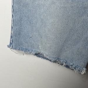 May include: Close-up of light blue denim shorts with a frayed hem. The shorts show signs of wear and tear, with a faded appearance and some light discoloration. The fabric has a textured look.