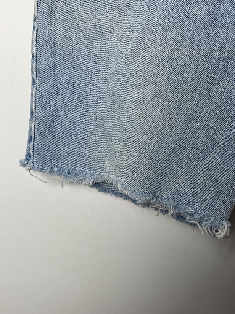 May include: Close-up of light blue denim shorts with a frayed hem. The shorts show signs of wear and tear, with a faded appearance and some light discoloration. The fabric has a textured look.
