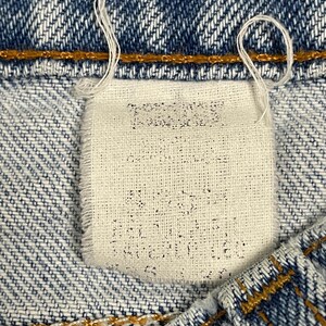 May include: Close-up of a denim jeans label. The label is rectangular, off-white, and stitched into the blue denim. Faint text is visible on the label. White threads hang from the label and the jeans' seam.