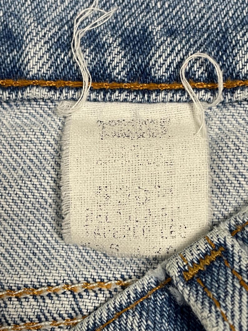 May include: Close-up of a denim jeans label. The label is rectangular, off-white, and stitched into the blue denim. Faint text is visible on the label. White threads hang from the label and the jeans' seam.