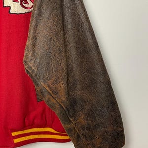 May include: Red and brown varsity jacket with the Kansas City Chiefs logo patch. The jacket has a red body with brown leather sleeves. The cuffs and waistband have yellow and red stripes.