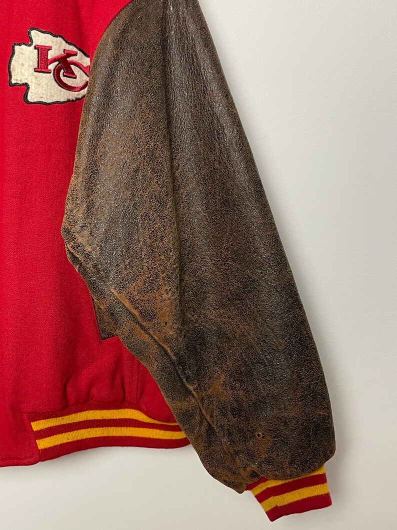 May include: Red and brown varsity jacket with the Kansas City Chiefs logo patch. The jacket has a red body with brown leather sleeves. The cuffs and waistband have yellow and red stripes.