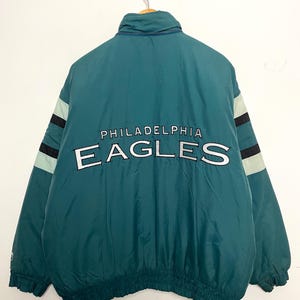 May include: Teal and black Philadelphia Eagles jacket with the team name and logo in white. The jacket has a high collar, a zipper closure, and elastic cuffs and hem. The sleeves feature black and light green stripes.