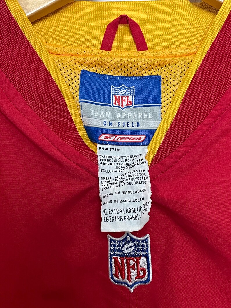 Puede incluir: Una chaqueta roja y amarilla de NFL Team Apparel con el logotipo de la NFL bordado en la parte delantera. La chaqueta tiene un cuello amarillo y un forro interior rojo. La etiqueta dice "XL Extra Large".