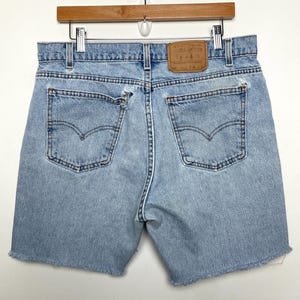 May include: Light blue denim shorts with a frayed hem, featuring two back pockets with the Levi's signature arcuate stitching. The shorts have a faded appearance and a leather patch on the back waistband. The shorts are hanging on a wooden hanger.