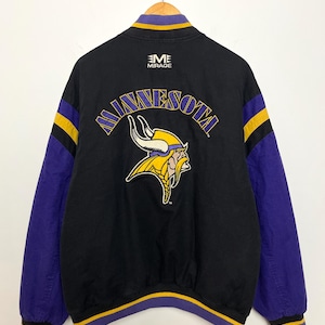 Puede incluir: Chaqueta negra con mangas moradas y detalles amarillos. La parte trasera presenta la palabra "MINNESOTA" en morado, junto con un logotipo vikingo amarillo y blanco. La chaqueta tiene cuello y puños acanalados con rayas amarillas y moradas.