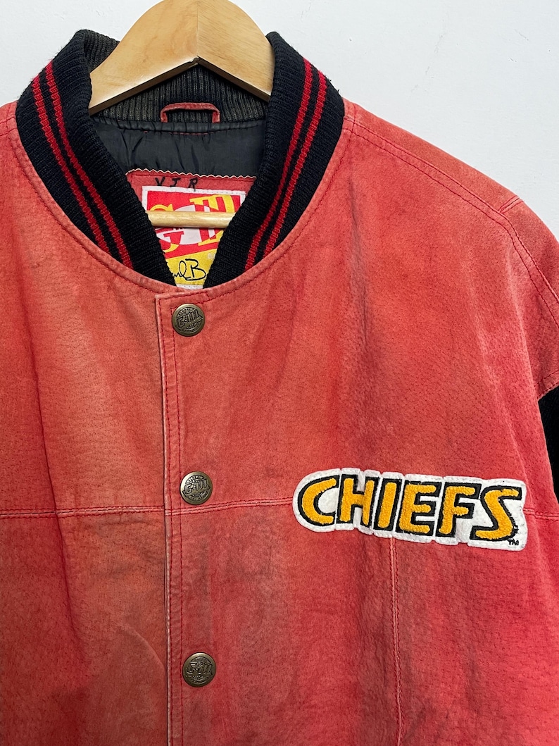 Puede incluir: Chaqueta de b&eacute;isbol de cuero rojo con ribetes negros y rojos alrededor del cuello y las mu&ntilde;ecas. La chaqueta tiene un gran parche amarillo y blanco "CHIEFS" en el lado derecho del pecho.