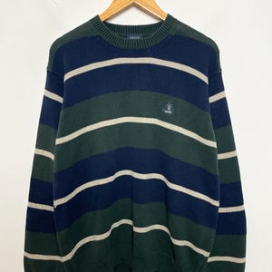 May include: A striped sweater in shades of navy, green, and white, hanging on a wooden hanger. The sweater features horizontal stripes and a small logo on the chest. The brand name "IZOD" is visible on the collar.