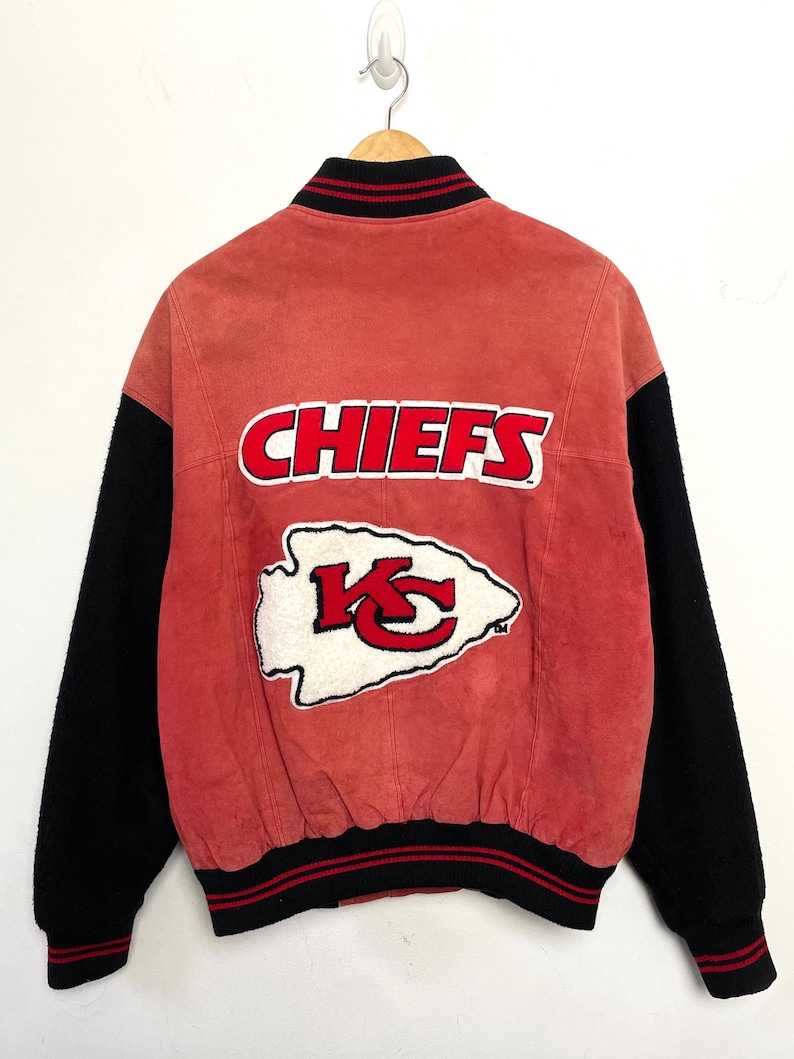 Puede incluir: Una chaqueta de b&eacute;isbol de gamuza roja y lana negra con el logotipo de los Kansas City Chiefs en la espalda. El logotipo es una punta de flecha blanca con las letras "KC" en rojo dentro. Las palabras "CHIEFS" est&aacute;n en letras blancas sobre un fondo rojo por encima de la punta de flecha.