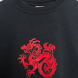 May include: Black crew neck t-shirt featuring a large red dragon design on the front. The dragon is stylized with intricate details. The shirt is hanging on a wooden hanger.