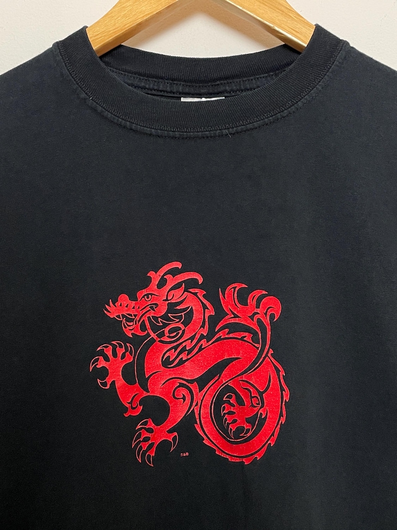 May include: Black crew neck t-shirt featuring a large red dragon design on the front. The dragon is stylized with intricate details. The shirt is hanging on a wooden hanger.
