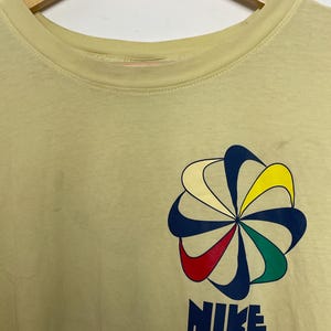 May include: Pale yellow t-shirt featuring a colourful floral design and the word "NIKE" in navy blue block letters. The floral design includes blue, yellow, red, and green accents. The shirt is hanging on a wooden hanger.
