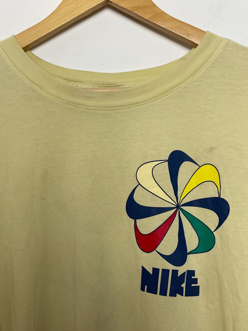 May include: Pale yellow t-shirt featuring a colourful floral design and the word "NIKE" in navy blue block letters. The floral design includes blue, yellow, red, and green accents. The shirt is hanging on a wooden hanger.