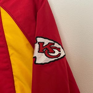 Puede incluir: Chaqueta roja y amarilla con un parche con el logotipo del equipo en la manga. El parche presenta una punta de flecha blanca con las letras "KC" en rojo. La chaqueta tiene un panel amarillo en la parte delantera y un cuerpo rojo.