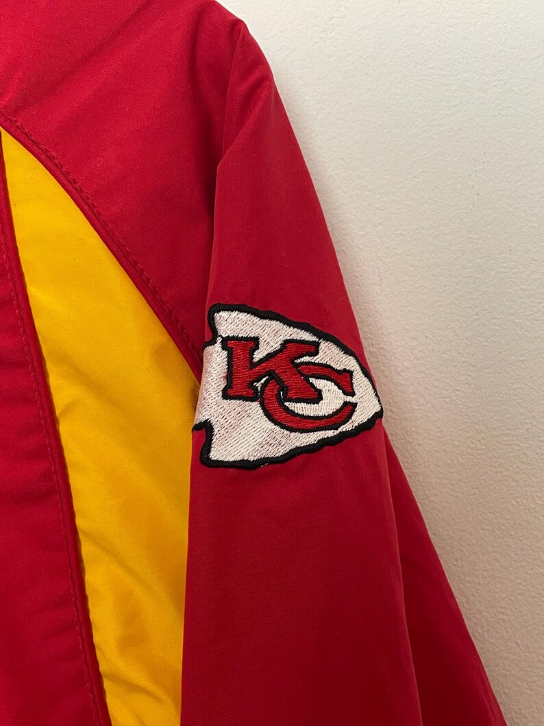 Puede incluir: Chaqueta roja y amarilla con un parche con el logotipo del equipo en la manga. El parche presenta una punta de flecha blanca con las letras "KC" en rojo. La chaqueta tiene un panel amarillo en la parte delantera y un cuerpo rojo.