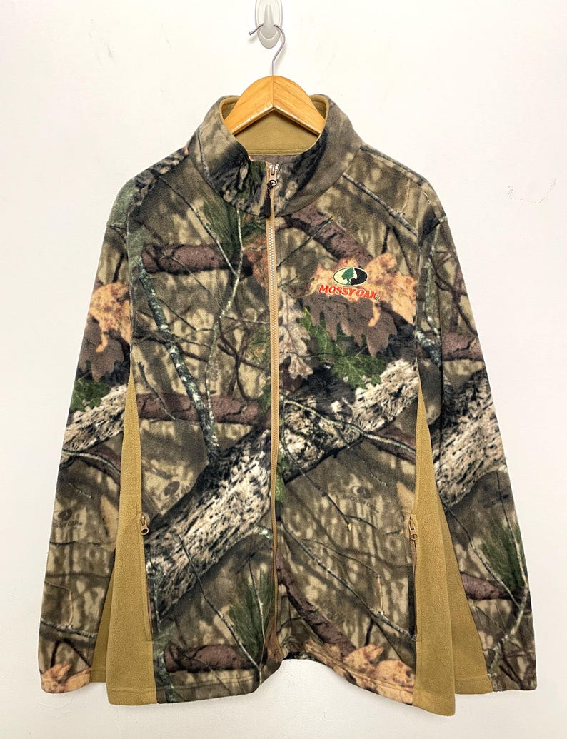May include: A full-zip fleece jacket with a camouflage pattern in shades of brown, green, and tan. The jacket features a stand-up collar, side pockets, and the "Mossy Oak" logo. The side panels are solid tan.