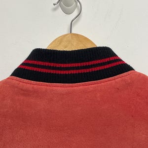 Puede incluir: Una chaqueta bomber roja con un cuello de rayas negras y rojas. La chaqueta est&aacute; colgada en una percha de madera.
