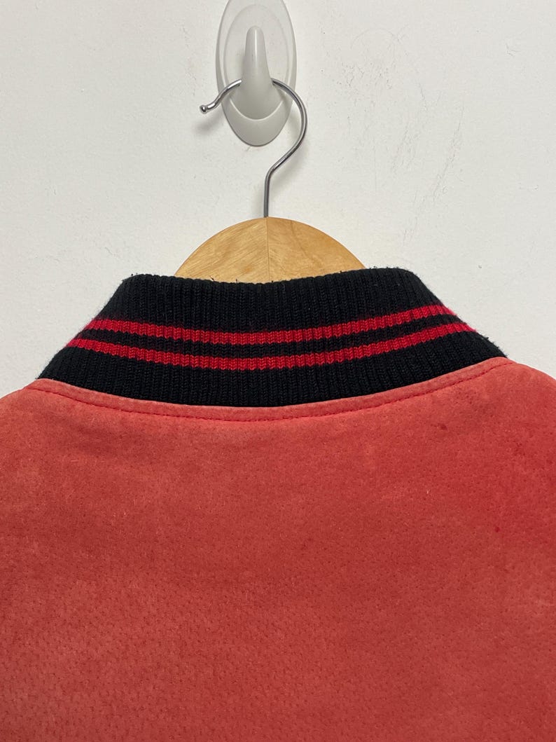 Puede incluir: Una chaqueta bomber roja con un cuello de rayas negras y rojas. La chaqueta est&aacute; colgada en una percha de madera.