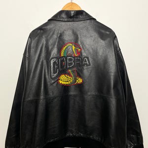 Op de afbeelding: Zwarte leren jas met een Cobra-logo geborduurd op de rug. Het logo heeft een geel, rood en groen cobra-ontwerp boven het woord "COBRA" in een vetgedrukte, gestikte letter. De jas heeft een klassieke bomberstijl met een zwarte geribde zoom en manchetten.